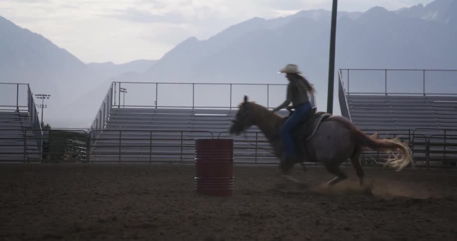 A barrel remains motionless on the sandy ground of the show arena as a horse with a female rider quickly runs around it as the sun is setting
