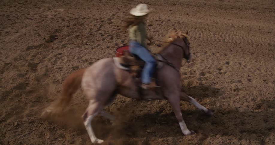 A cowgirl rides a horse quickly around a rodeo barrel in a dirt pen