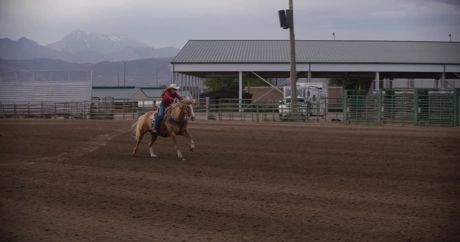 A cowgirl rides on the back of a horse on a dirt barrel racing track under a cloudy sky near the mountains