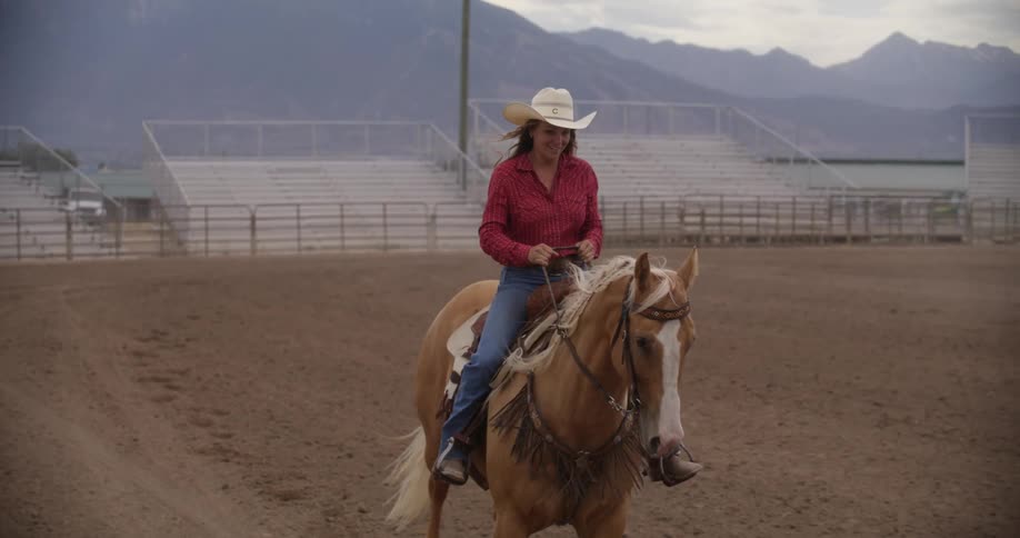 A woman dressed in horse riding attire wearing a cowboy hat smiles as she rides on the back of a light brown colt across a dirt horse racing track with empty bleachers and mountains behind her in a cloudy day