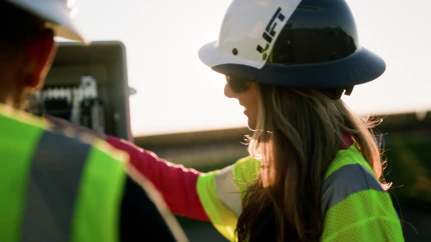 A woman and a man wearing hardhats and vests checking an electrical box outdoors during the day