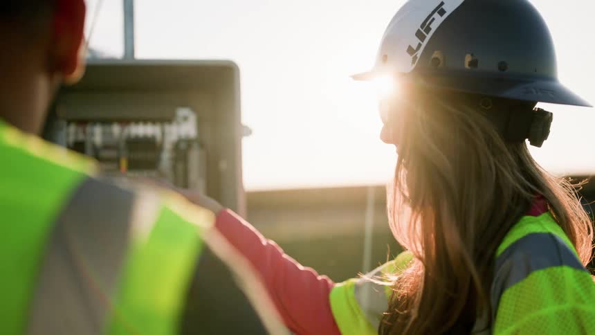 Two people wearing vests and hardhats checking an electricity box on a sunny day