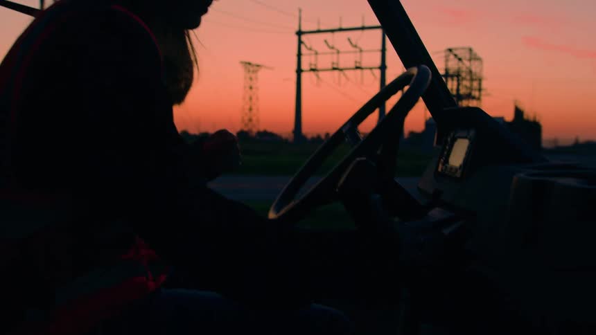 The silhouette of a man wearing an orange vest gets into a vehicle and shifts it into gear