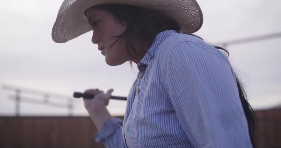 A female ranch hand wears a cowboy hat holds a horse whip moving it across her body from one side to the other behind her back looking forward as she starts walking inside a fenced in pen on a ranch under a cloudy sky as the sunsets