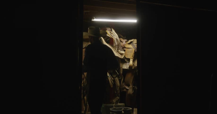 Cowboy carrying rope walks into shed holding horsetack