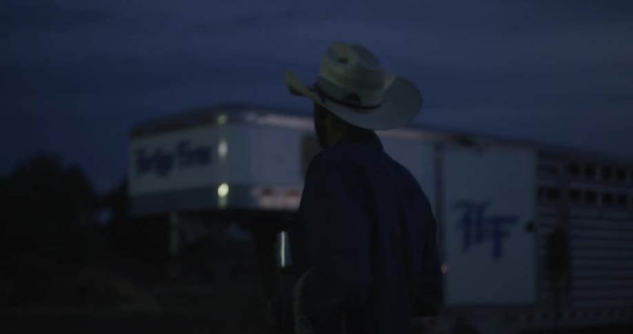 A man in a cowboy hat holds a cup and walks past a horse trailer at sunset
