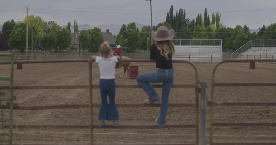 Two girls on a fence watch a woman ride a horse in an enclosure
