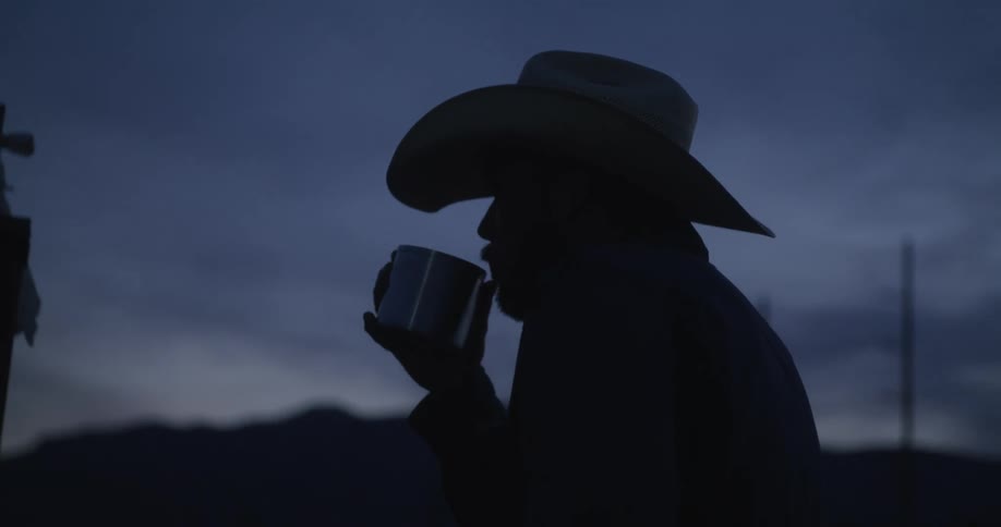 A man in a cowboy hat stands outside and drinks from a cup at sunset