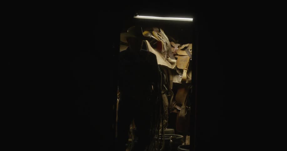 Cowboy carries horse tack from shed at night