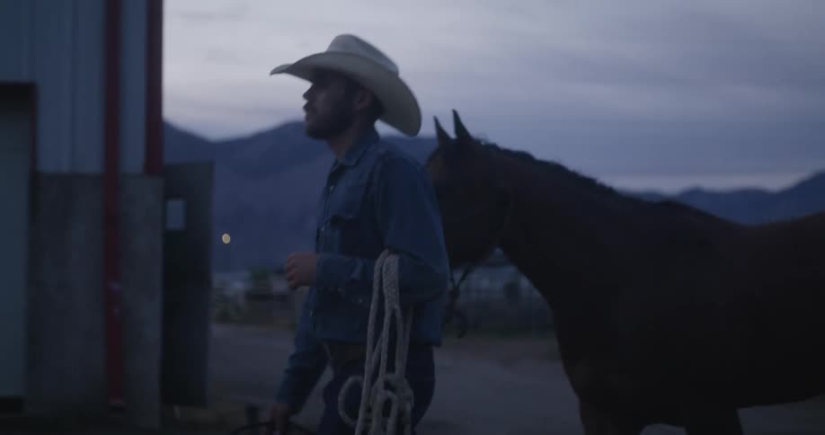 A man in a cowboy hat and with ropes in his hand is walking with a horse behind him