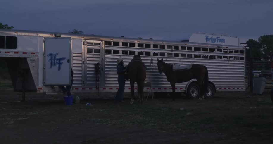 Person saddle up a horse tied to a parked trailer at sunset