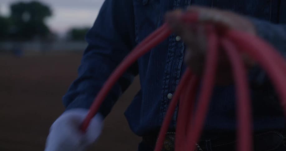 Cowboy stands in rodeo arena with his horse and prepares his rope
