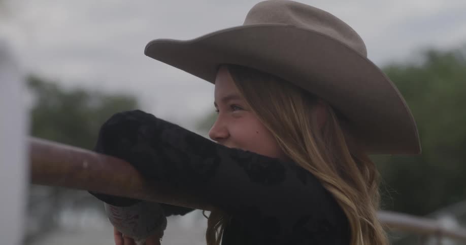 A little blonde girl in a cowboy hat smiling and with her arms on a fence
