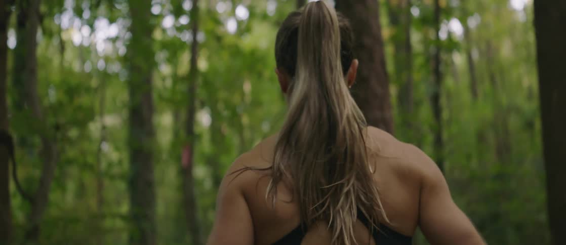 A womans blonde ponytail swings back and forth across her upper back and bare shoulders as she jogs through a wooded area in the daytime