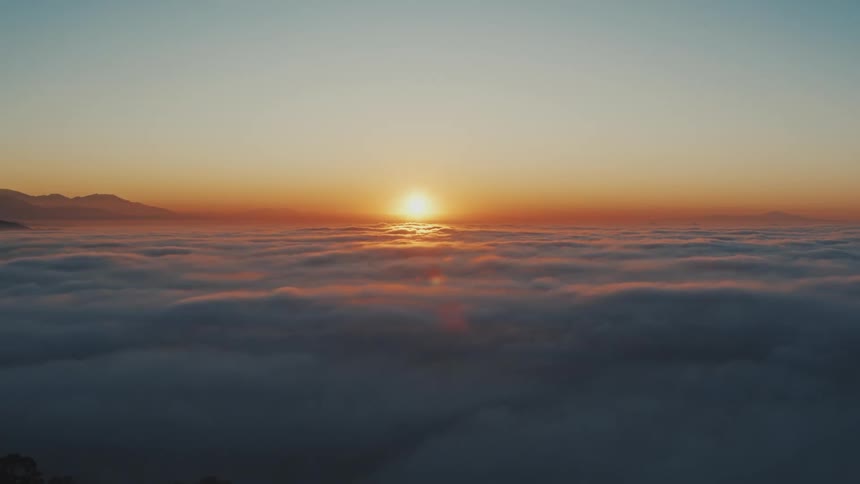 View high above the clouds as the sun glows on the horizon among mountaintops