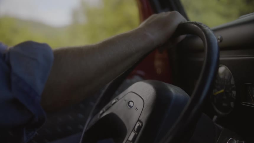 A person drives on uneven terrain with on arm stretched out grasping the top of the steering wheel periodically turning it slightly to straighten out the vehicle while resting the arm on the door window
