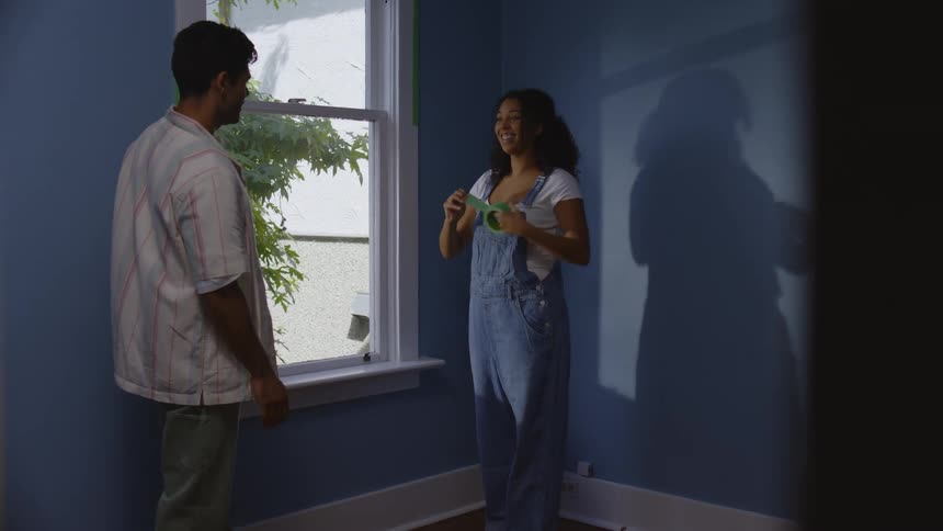 A couple stands in an empty room with blue walls and puts tape along a window frame