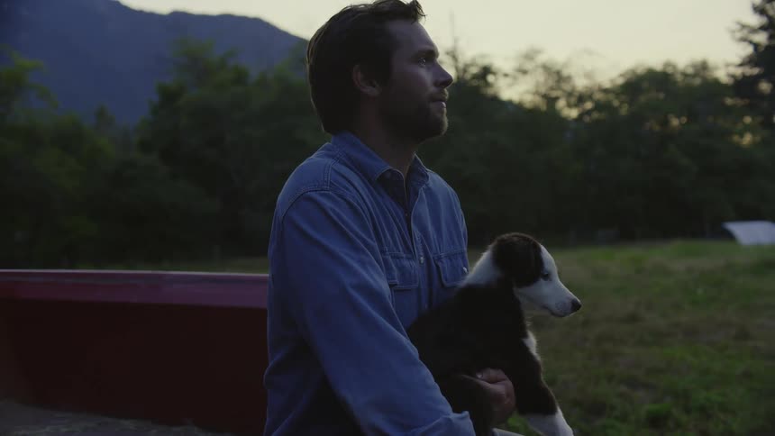 Man sitting on the tailgate of his red pickup truck holding a dog on his lap at sunset