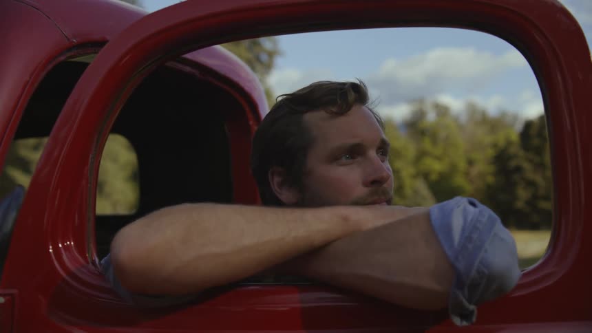 A man turns his head which is resting on his arms that are rested in the open window of the drivers door of an old red truck