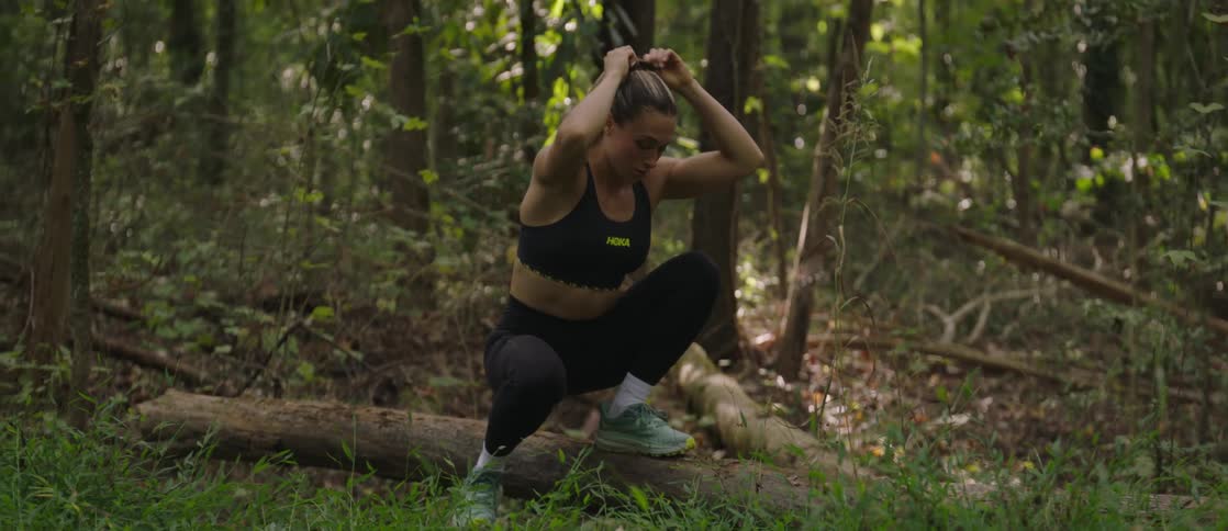 A woman in running attire breathes heavily andpulls back her hair as she rests squatted down with one foot on a log and the other on the ground in the middle of a wooded area