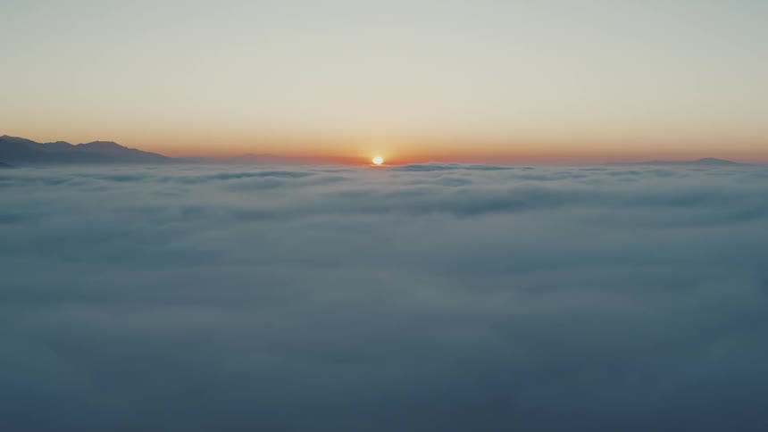 View high above the clouds as the sun glows on the horizon among mountaintops