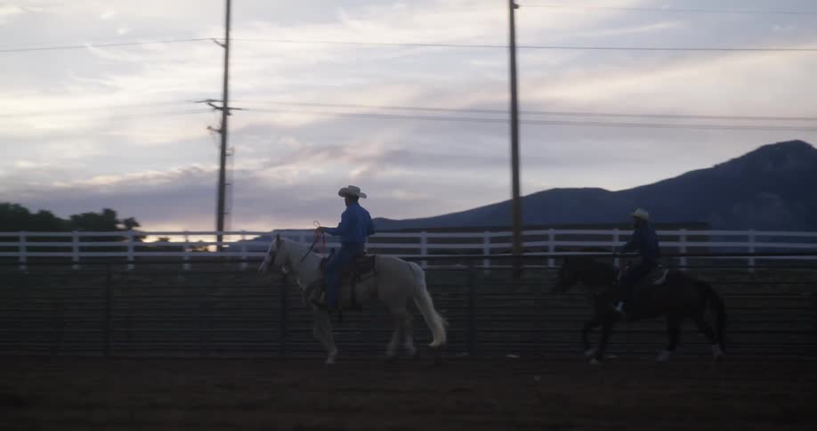 Two horsemen ride horses through pasture on ranch near mountains after dusk