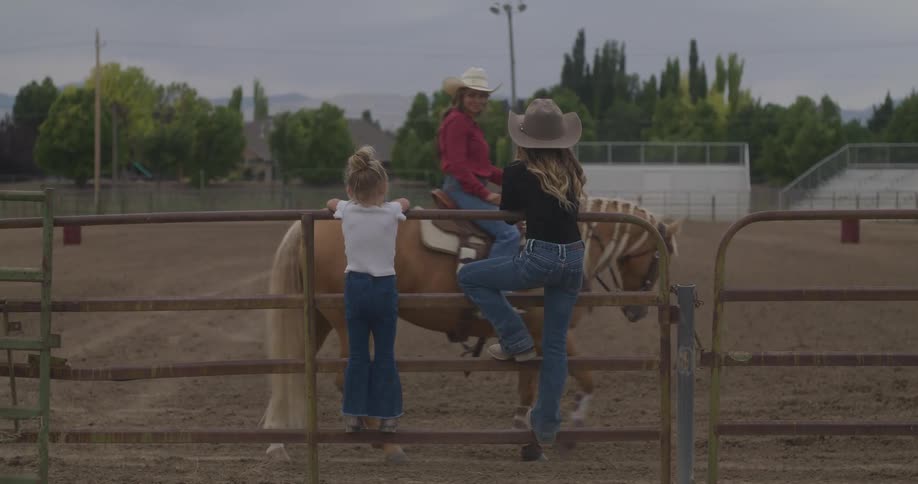 Woman and a girl in the grass of a rodeo arena with a cowgirl riding a horse passing by
