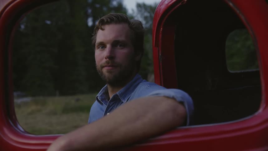 A portrait shot of a blonde male farmer smiling while his arm is in the window of the parked red truck