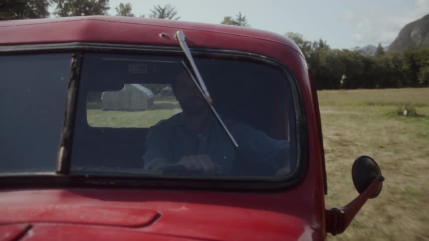 A man driving a red pickup truck on a grass field during the day
