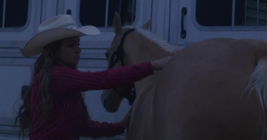 A woman in a cowboy hat brushes a horse next to a trailer