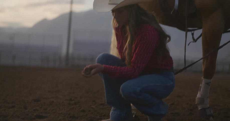 Portrait of a cowgirl next to her horse crouching and grabbing the ground with her hand