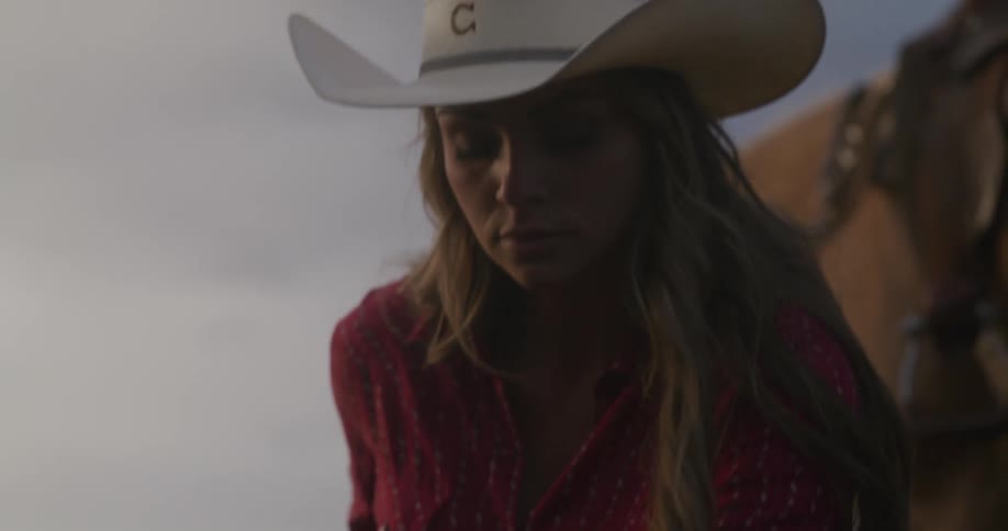 A woman dressed in cowgirl attire and a white cowgirl is bent down picking up the dirt and feeling it with one hand with her horse behind her before turning her head towards the setting sun
