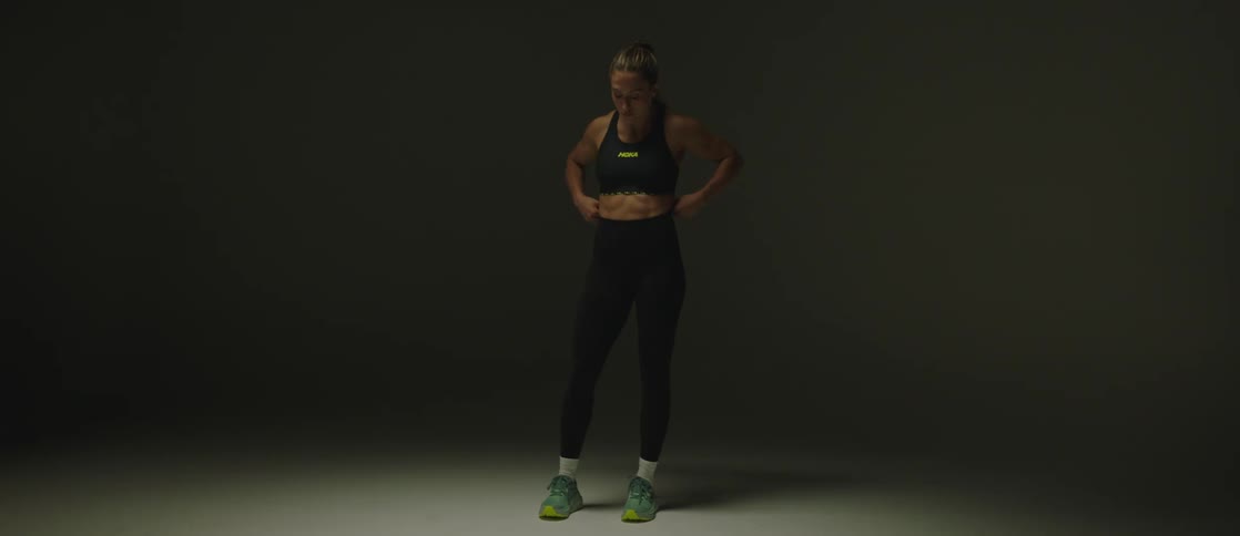 A woman wearing a sports bra and running pants with tennis shoes steps out of the darkness and stands with her hand on her hip