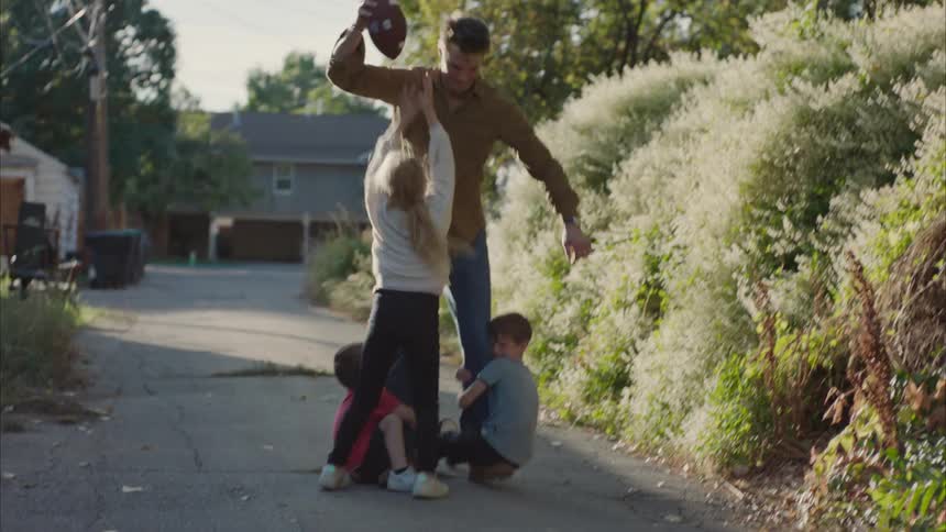 Father holding an american football with his two sons clinging to his leg and his daughter jumping trying to catch the ball