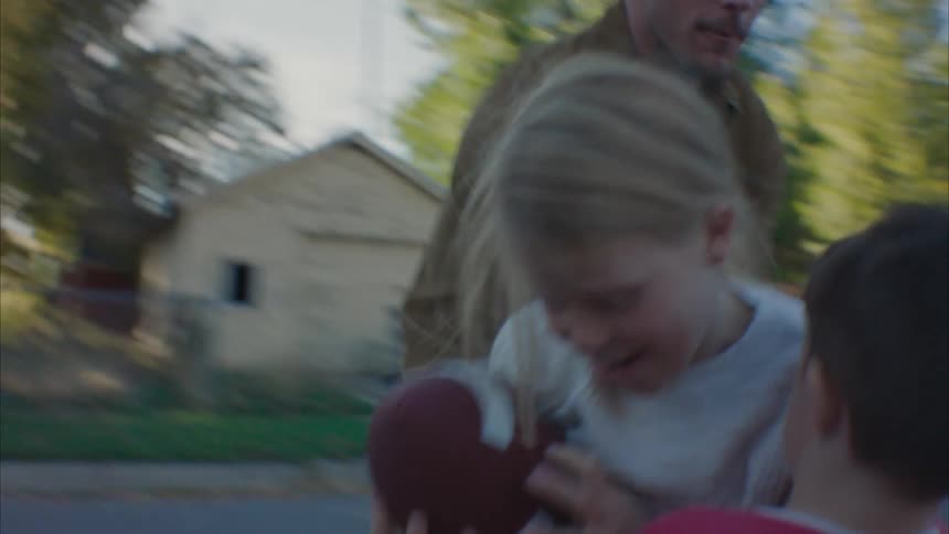 Girl running with an american football on a neighborhood street with her father and siblings trying to catch her