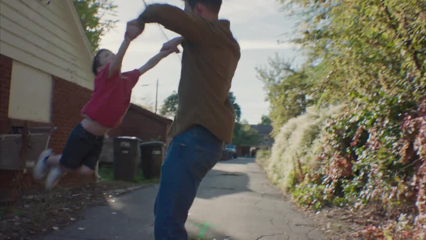 A man is standing in an neighborhood alley swinging his young son around by his hands