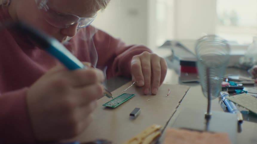 A little blonde girl wearing glasses is soldering a piece of hardware
