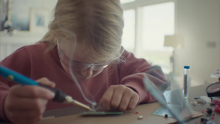 A little blonde girl with glasses is soldering an electronic part with a soldering iron