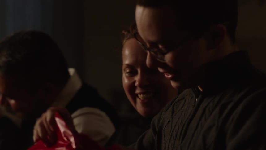 A present wrapped in red paper is being opened by a teenage boy while his mom watches happily beside him and a male and younger boy interact beside her with everyone dressed in nice sweaters