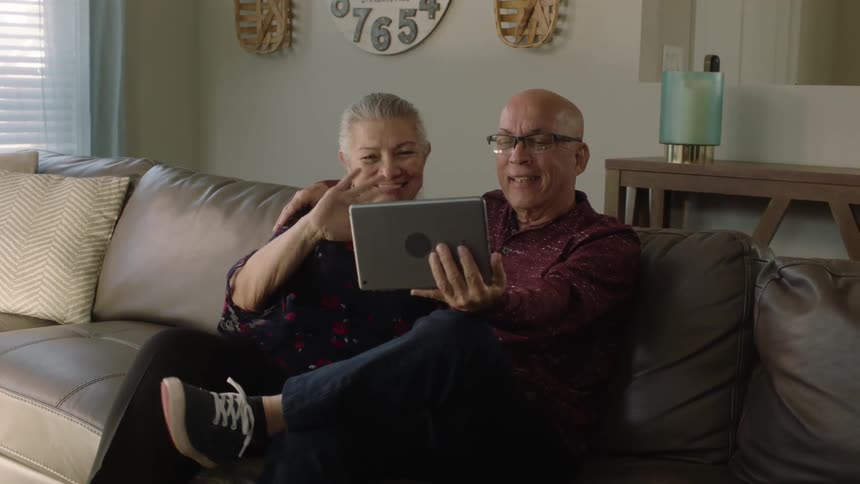 An elderly couple sitting together on the couch smile as they wave into a tablet computer