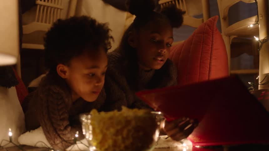 Two children are reading something in a makeshift hut made of sheets and pillows and some lights
