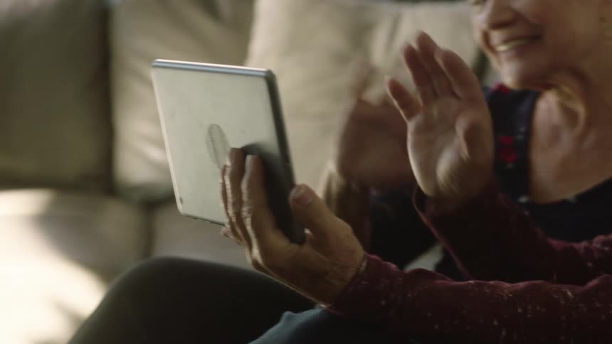 An elderly man and woman sitting on a couch beside each other wave at the screen of a tablet being held by the man in front of them