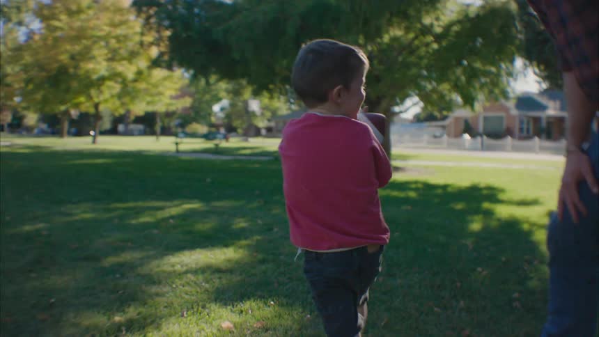 A little boy runs in his yard with a football as his father runs and catches him