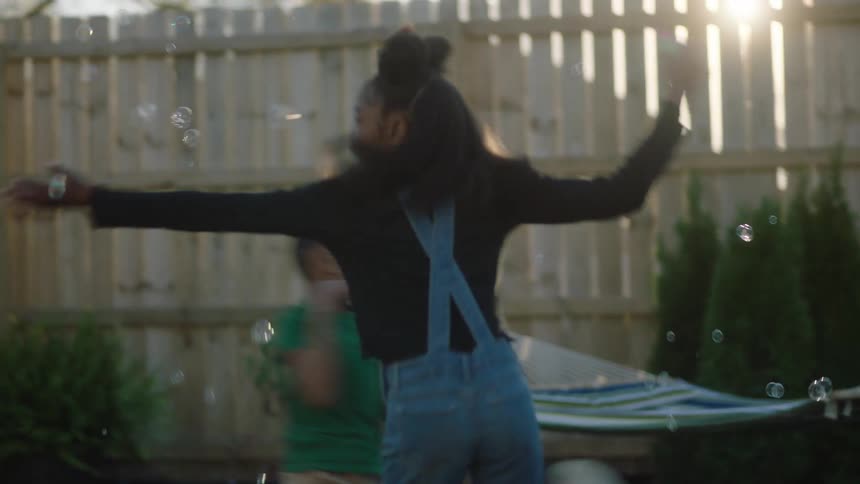 An older female child spinning with a bubble wand and younger male child running around her popping the bubbles in a fenced in yard as the sun is setting