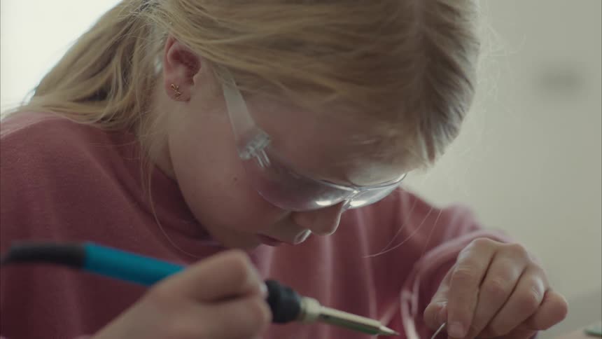 A little girl is soldering something while wearing clear glasses