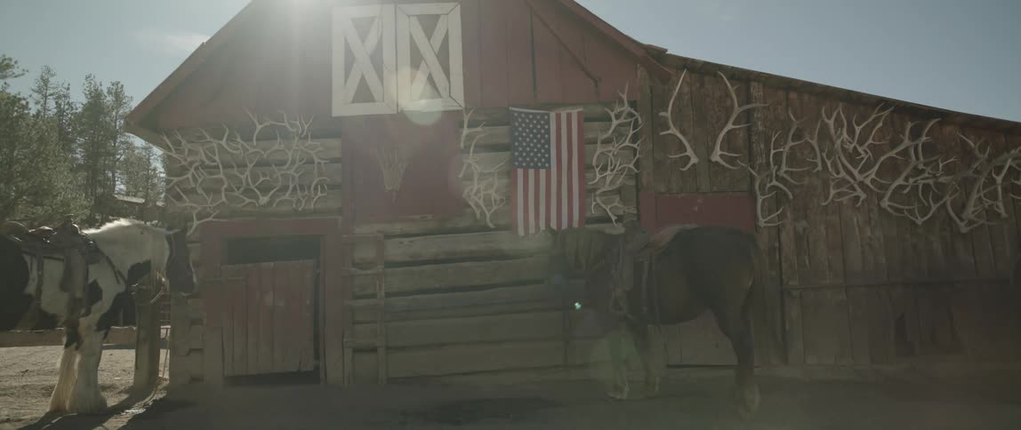 A row of antlers and an american flag hang on the outside facade of a red wooden barn with horses tied up shadowed from the sun beaming down from a clear blue sky above