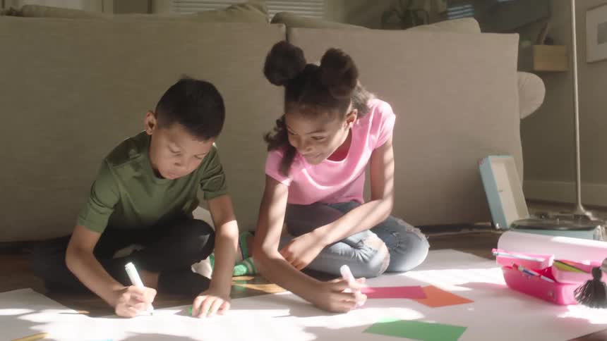 Boy and girl writes on a long white paper while seated on the living room floor at midday