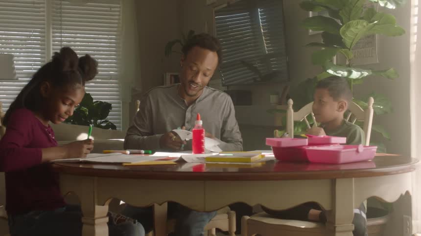 Man helps his children with their homework while they converse sitting at a round table indoor at midday