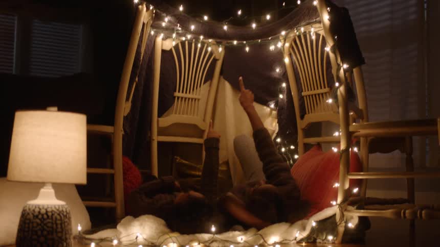 Two girls lying in an improvised fort made with chairs and christmas lights in the room at night