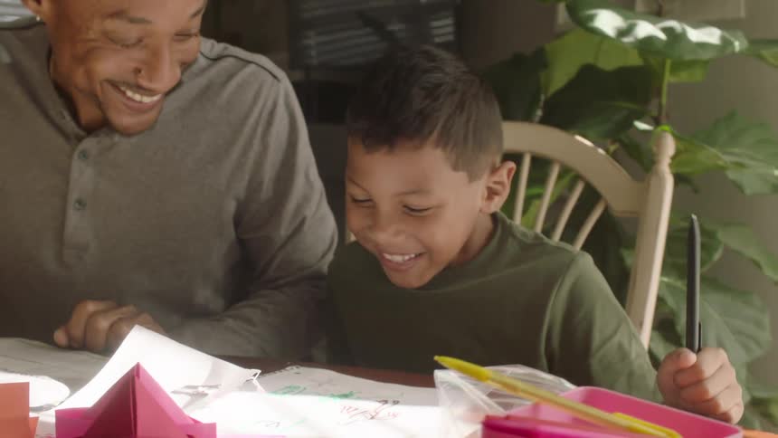 Man and boy sit at table doing paper craft indoors during the day